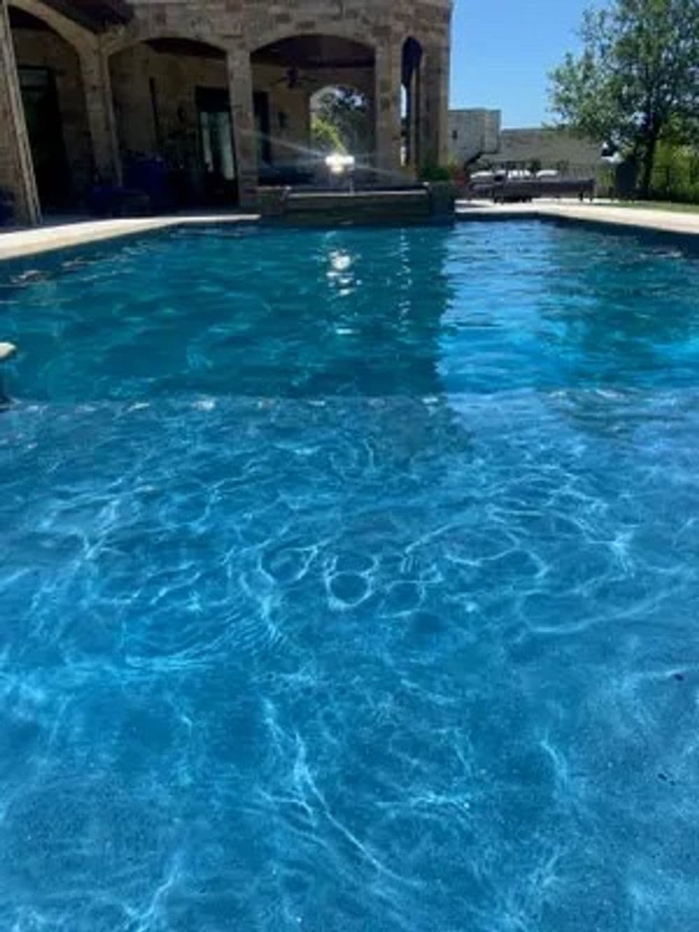 Crystal-clear swimming pool water reflecting sunlight, surrounded by a stone patio and greenery.