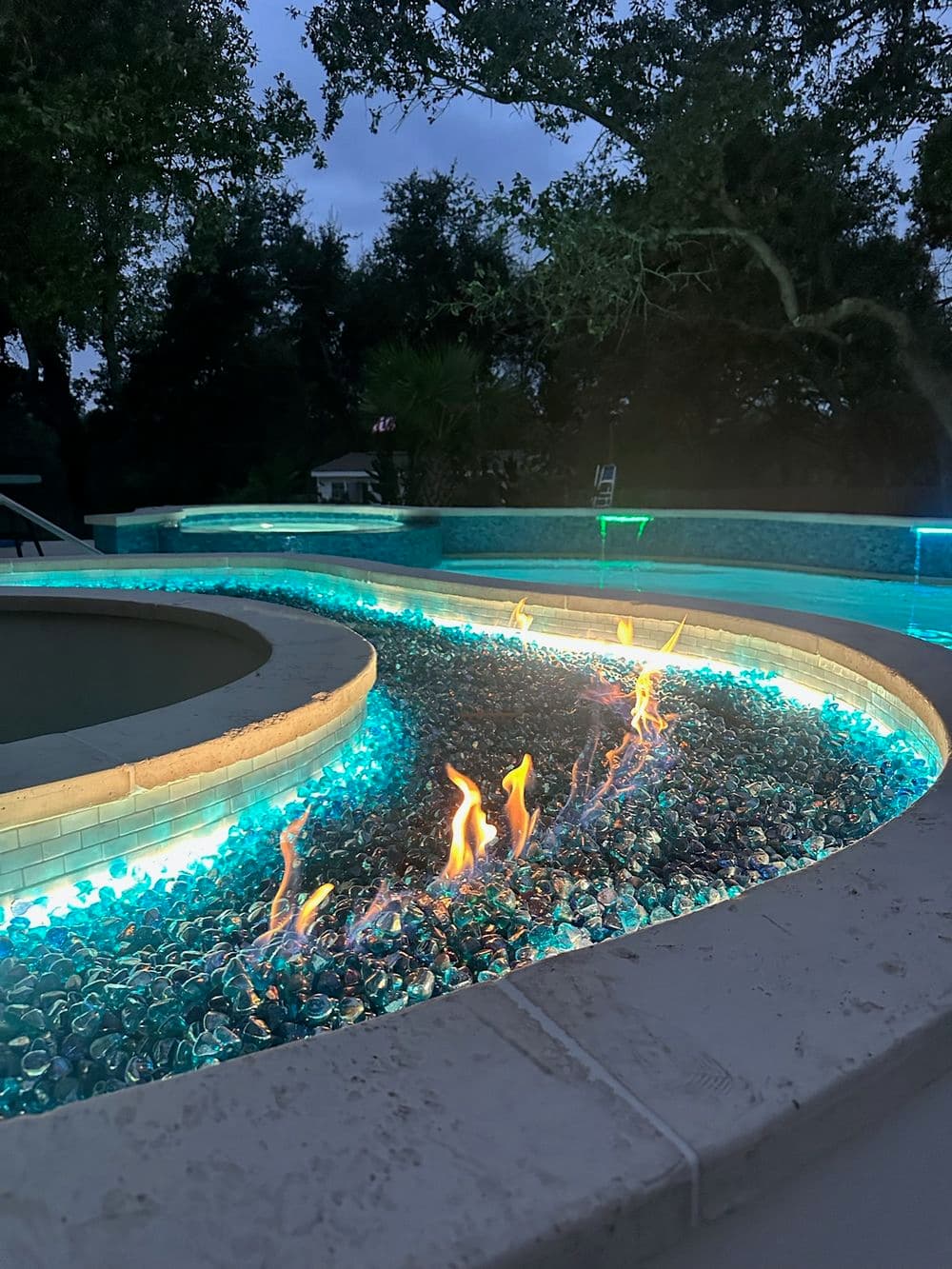 Modern outdoor pool with glowing blue glass stones and flame features at dusk.