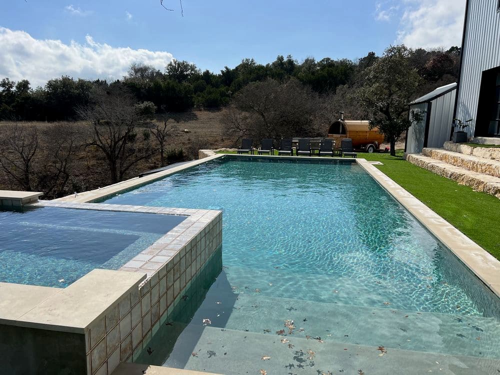 Modern infinity pool with spa, surrounded by landscaping and wooded area. Clear blue water.