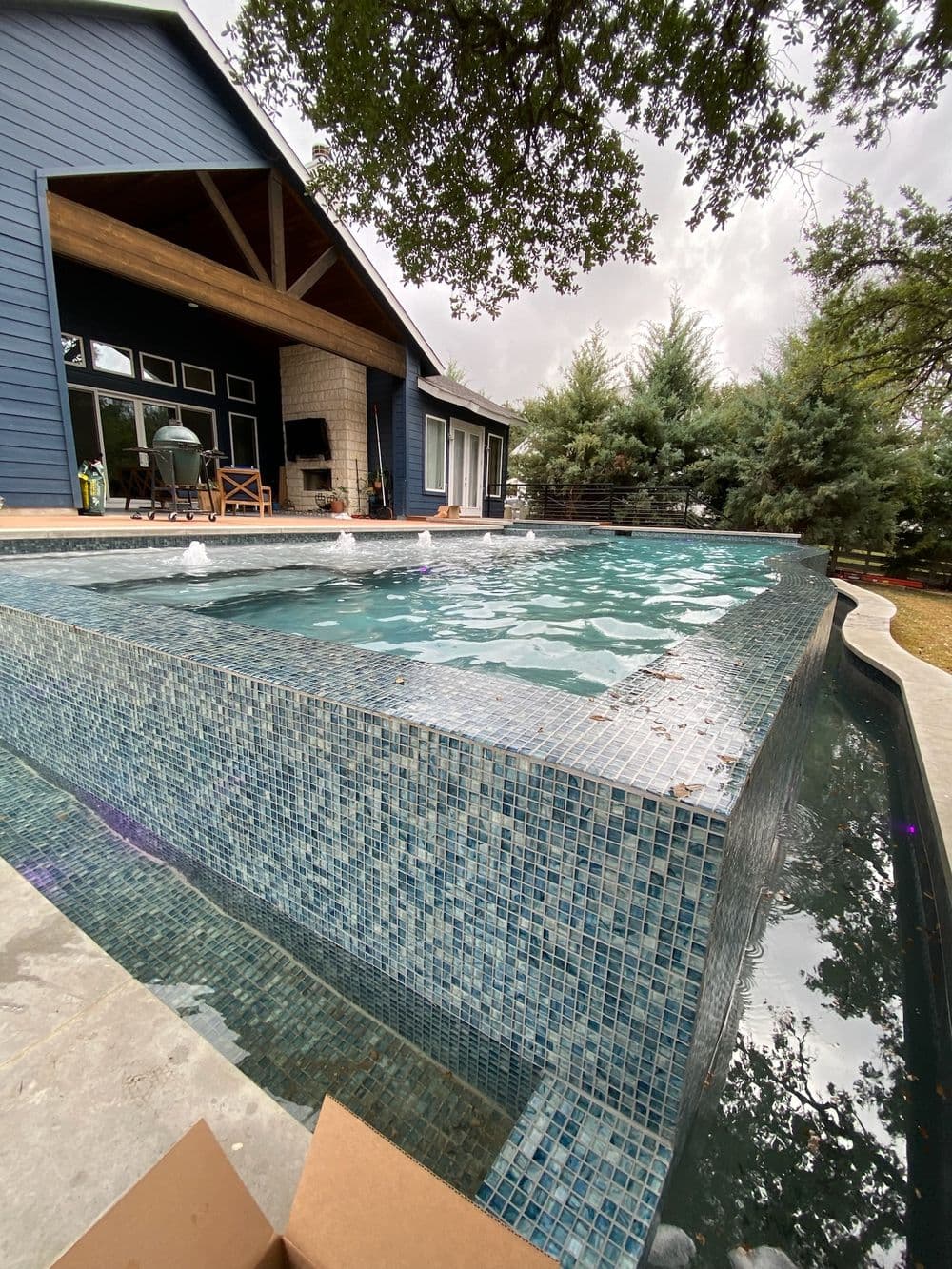 Modern backyard pool with mosaic tiles, surrounded by trees and a blue house. Relaxing oasis.
