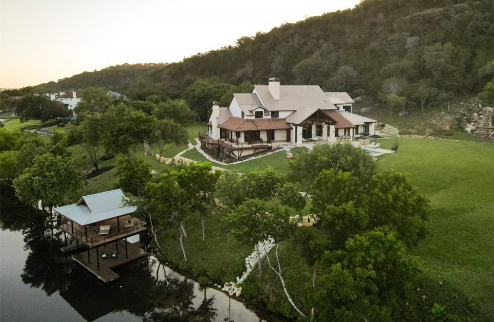 Aerial view of a luxurious riverfront home with a spacious lawn and dock amidst lush greenery.