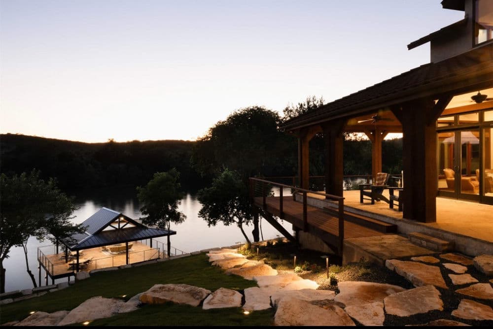 Elegant riverside home at sunset with a deck, stone landscaping, and a boathouse.