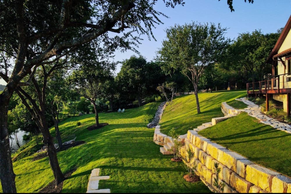 Lush landscaped yard with stone wall, trees, and clear blue sky next to a serene lake.