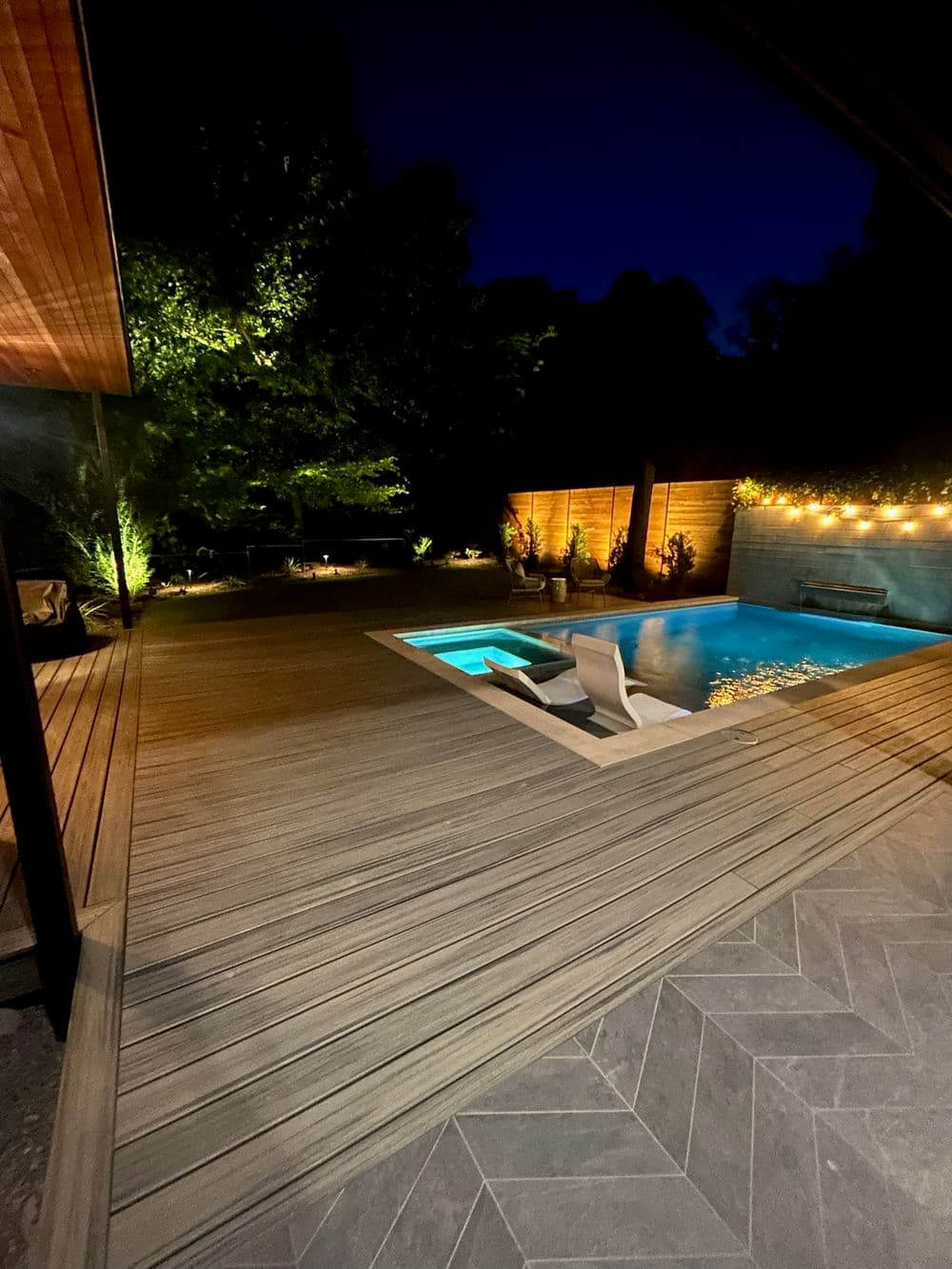 Modern backyard at night featuring a pool, wooden deck, and illuminated landscaping.