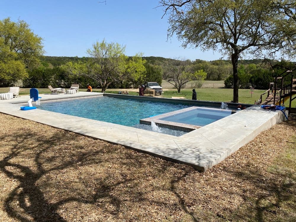 Outdoor swimming pool with spa surrounded by trees and grassy area, perfect for summer relaxation.