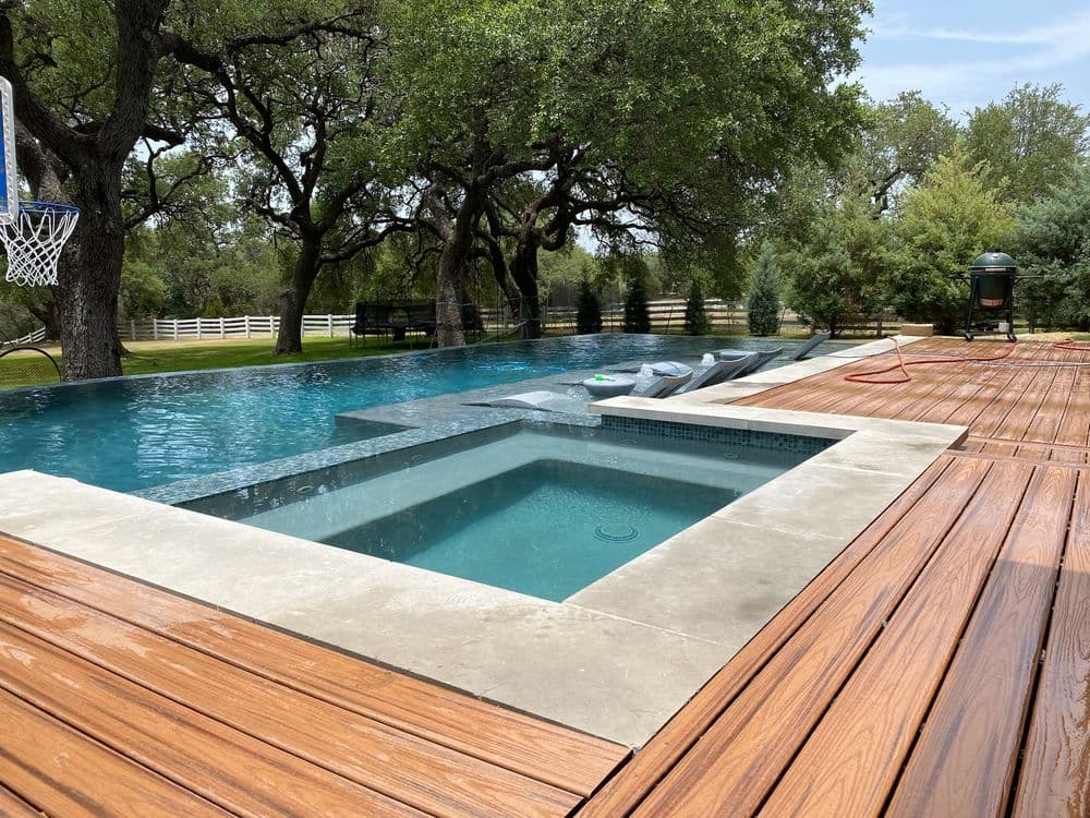 Modern backyard pool with spa, wooden deck, and landscaped greenery under blue sky.