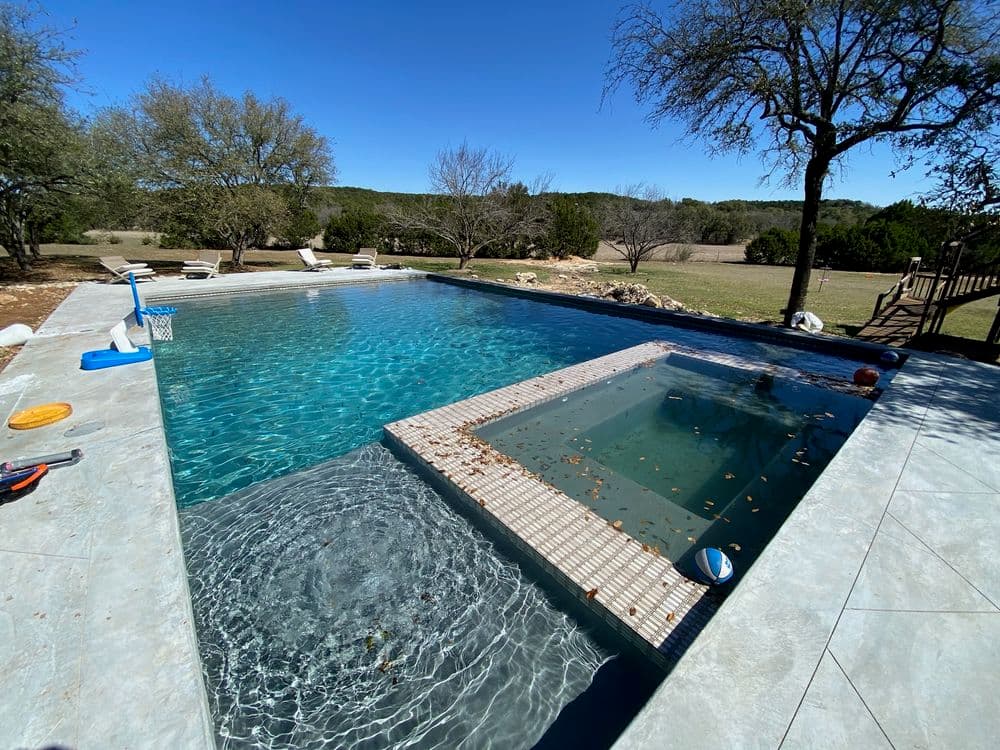 Luxury backyard swimming pool with hot tub, surrounded by trees and clear blue sky.