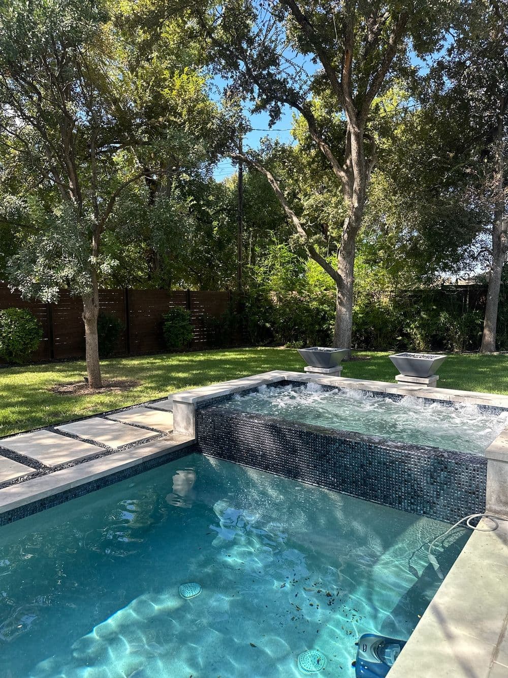 Luxury backyard pool with spa, surrounded by lush greenery and sunny skies. Relaxing oasis.