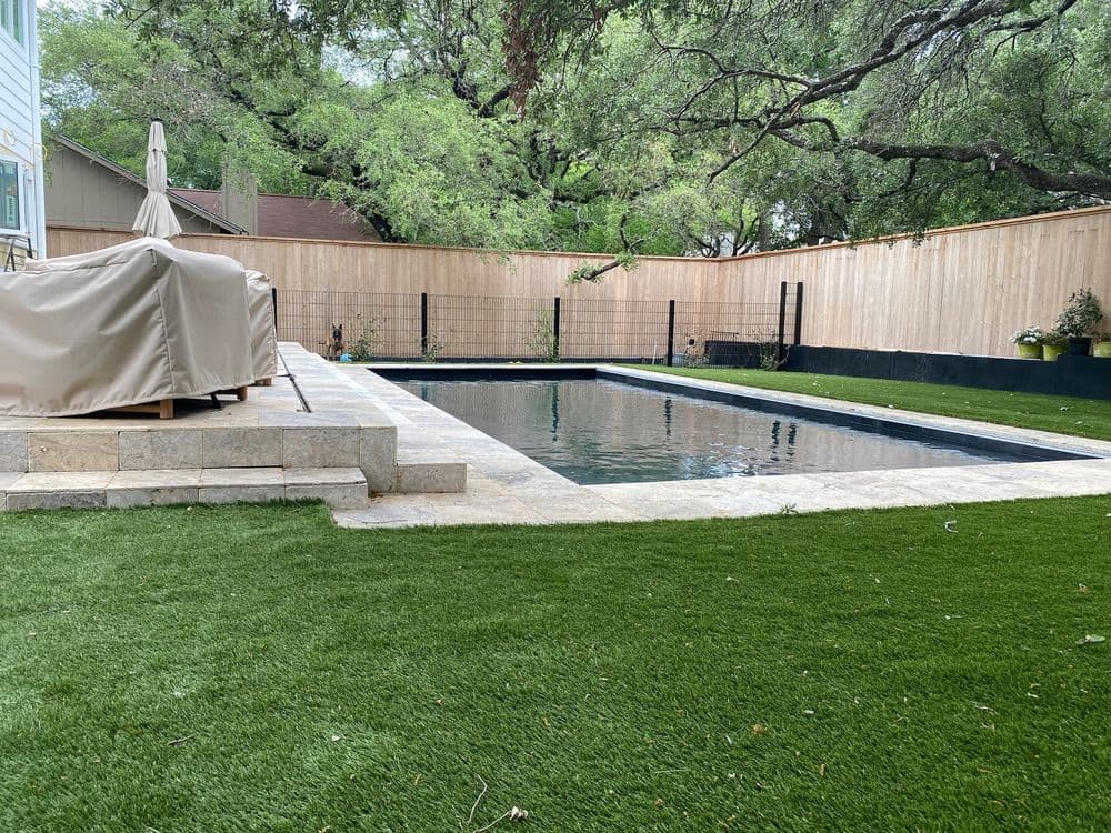 Backyard pool with stone patio, surrounded by artificial grass and wooden fence.