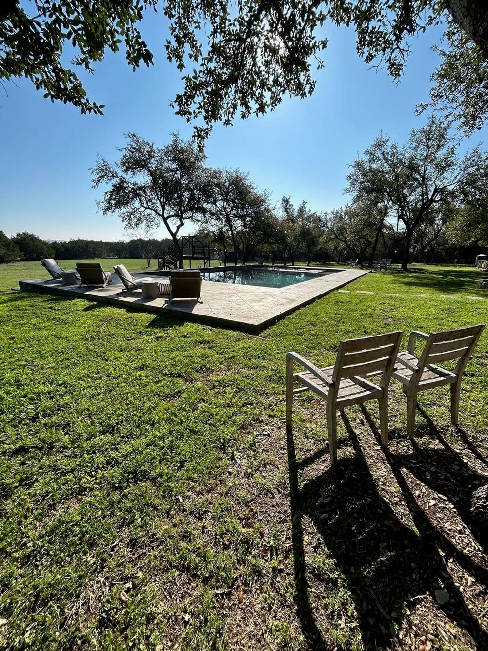 Sunny outdoor pool area with lounge chairs and trees, surrounded by lush green grass.