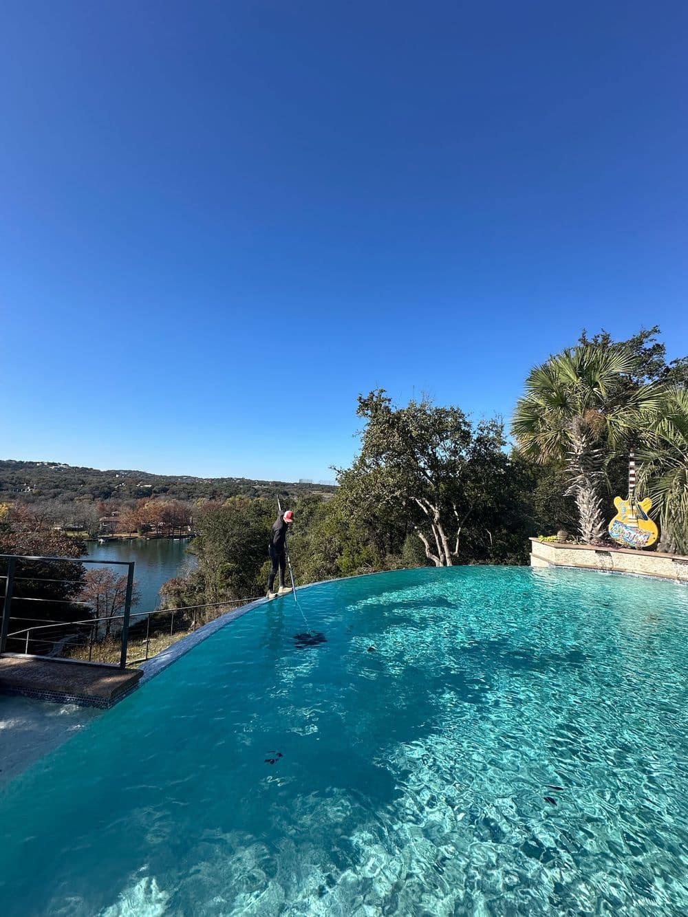 Person cleaning an infinity pool with a clear blue sky and scenic landscape in the background.