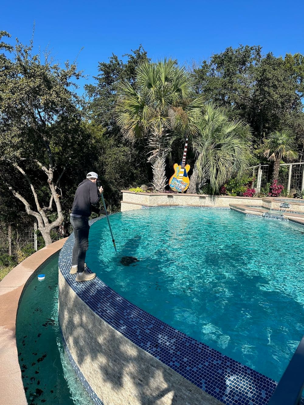Person cleaning a pool with a skimmer, surrounded by palm trees and vibrant landscaping.